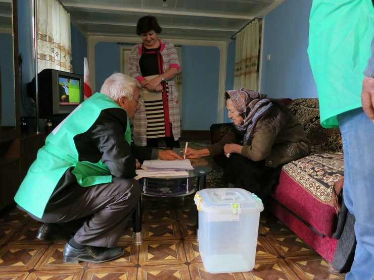 An older woman and a man lean over the hood of a dark blue car. The man is explaining the ballot to the woman. Behind them is a mobile ballot box and a team that moves the box.