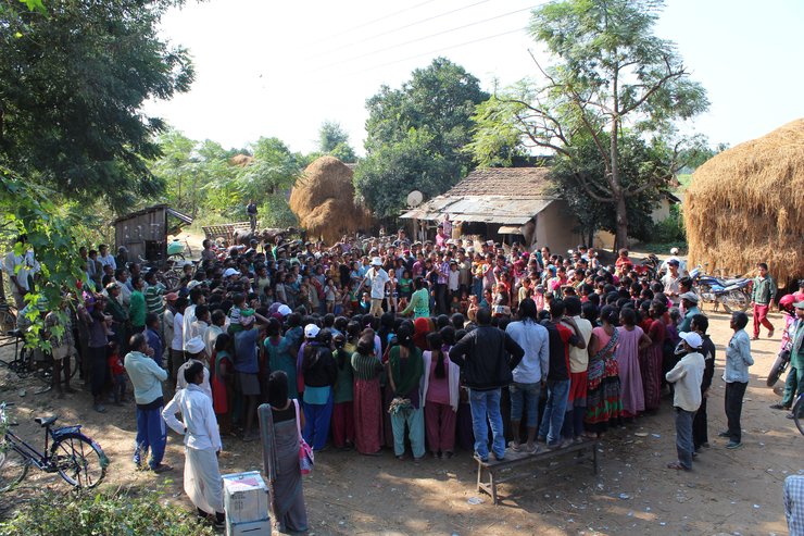 About 60 to 70 people are standing in a circle outdoors. There are trees and piles of straw in the far background. In the middle of the circle there appear to be two people (a man and a woman) who are talking to each other.  