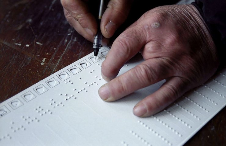 A sheet of long paper with Braille and holes to mark ballots sits on top of a desk. Two hands are poised to fill it out; the left hand appears to have four fingers.