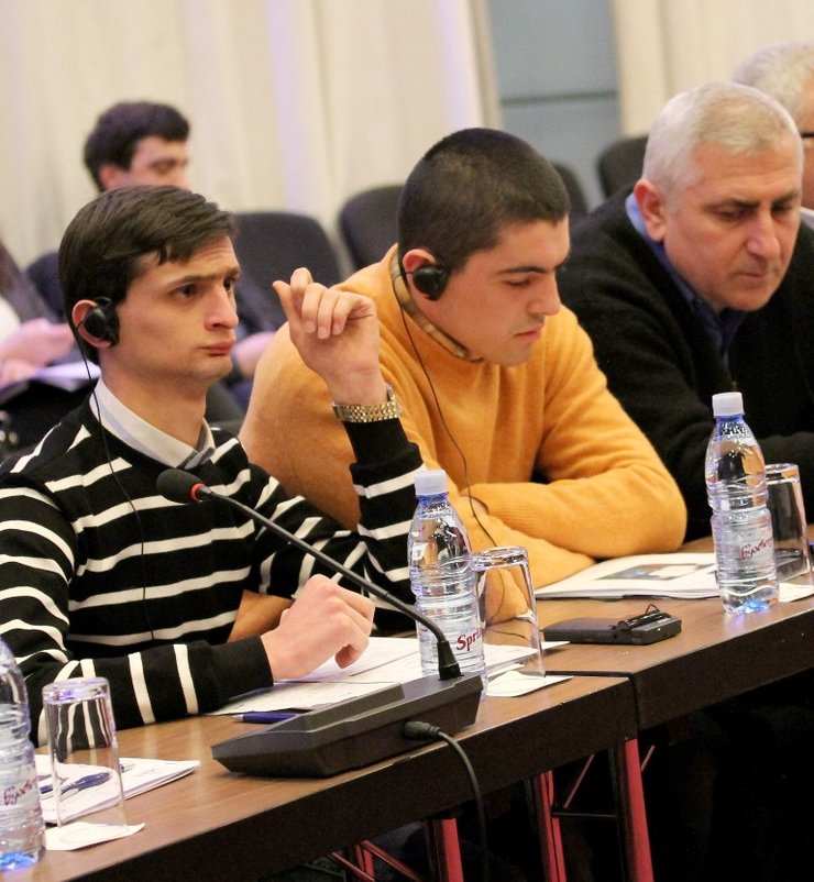 In the foreground is a table. On the left is a man sitting in a black and white striped shirt, listening to headphones. On his left is a man in a yellow shirt also listening to headphones.