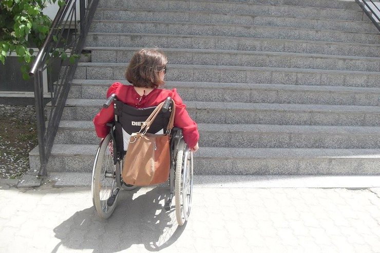 A woman using a wheelchair and wearing a red shirt waits in front of a large staircase.