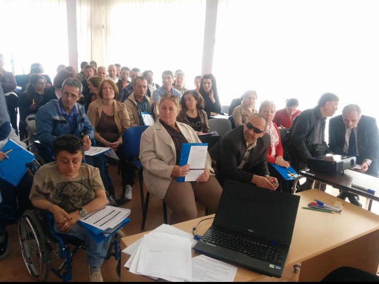 In a large room, there are about 30-40 poepl. In the front row are two people (a man and a woman) using a wheelchair.